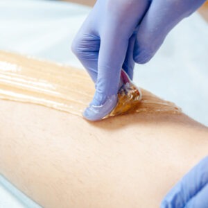 Eine Hand mit blauen Handschuhen trägt eine Wachsschicht auf einem Bein auf, während eine Folie abgezogen wird.