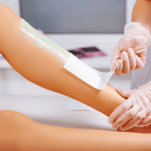 Eine Person mit Handschuhen entfernt Wachs von einem Oberschenkel, während eine andere Beinform im Hintergrund zu sehen ist.